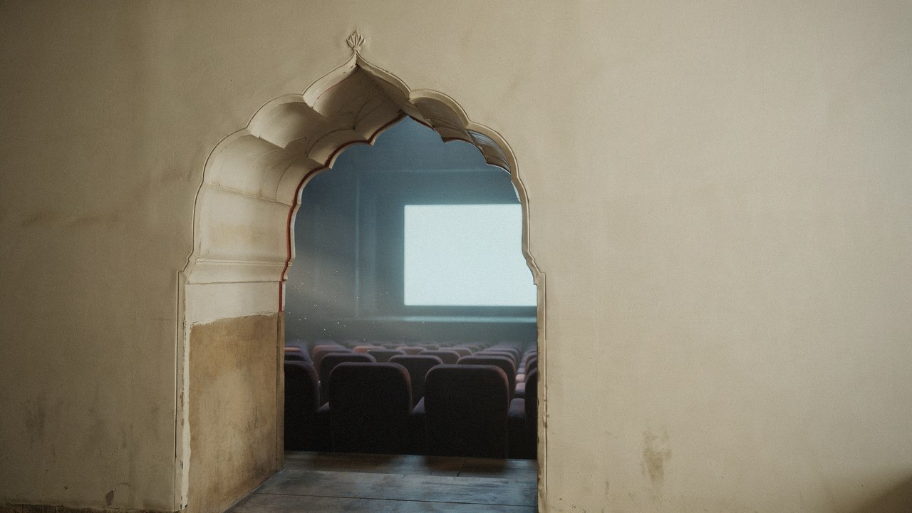 A pale Mughal-style cusped stone archway in a cream plaster wall, opening onto a dark cinema auditorium of empty velvet seats facing one bright blank screen — the fourth wall, framed. Cinema Kept Breaking the Wrong Wall by Hana Mattar, Bombay Blanc.